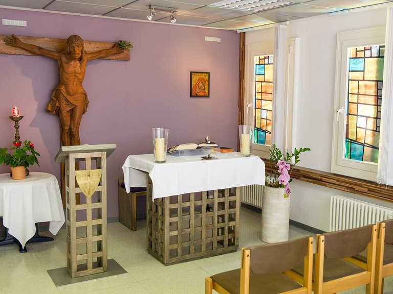 Innanansicht: moderne kleine Kapelle mit Holzkreuz, Osterkerze, kleinem Altar und einer Stuhlreihe